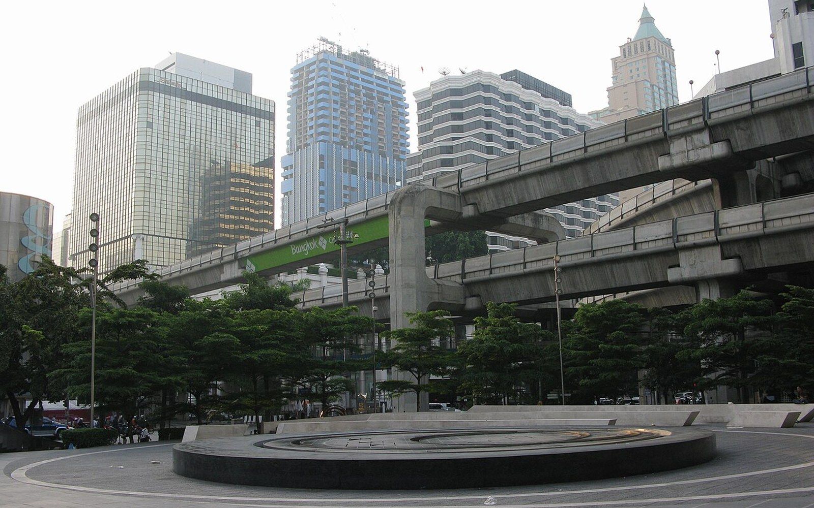 Elevated skytrain viaduct curves above a plaza, with high-rise towers in the background